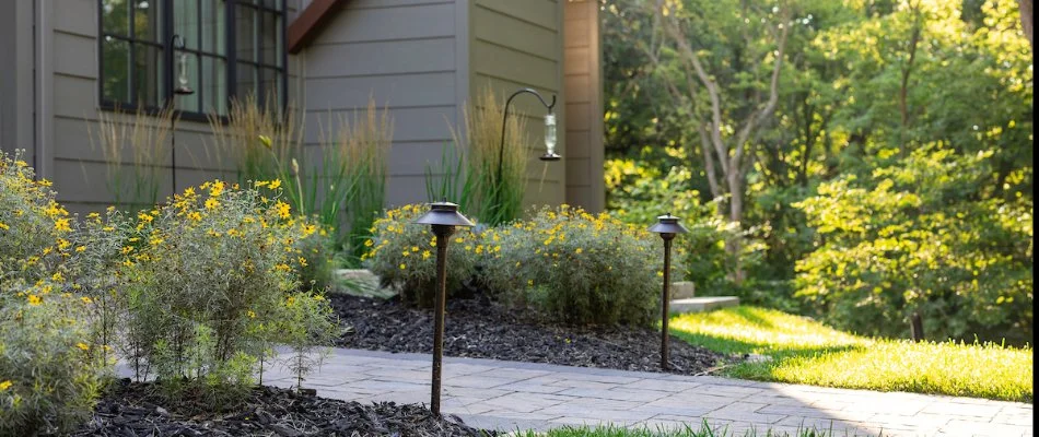 Inviting garden path lined with decorative lighting and vibrant yellow flowers, adjacent to a charming home.